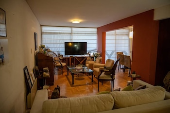 Una sala de estar y comedor de aspecto residencial, con muebles de madera, sofá claro, televisión, y persianas blancas en las ventanas