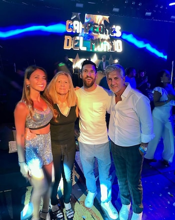 Lionel Messi, Antonela Roccuzzo, Jorge Messi y Celia Cuccittini sonríen en un evento festivo con luces azules y un cartel 'Campeones del Mundo' de fondo