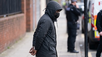 Un hombre con las manos esposadas a la espalda viste un abrigo negro con capucha y mascarilla negra junto a agentes de policía.