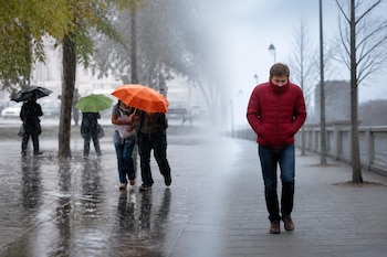 Peatones con paraguas caminan bajo la lluvia a la izquierda y un hombre con chaqueta roja camina por una acera seca a la derecha.
