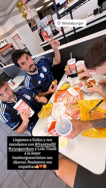Tres personas en una mesa de un restaurante. Hay hamburguesas, papas fritas, bebidas en vasos Whataburger y salsas. Dos hombres usan camisetas de Argentina