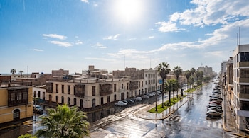 Edificios de estilo tradicional y palmeras flanquean una avenida con calles mojadas bajo la lluvia y la luz directa del sol en el cielo.