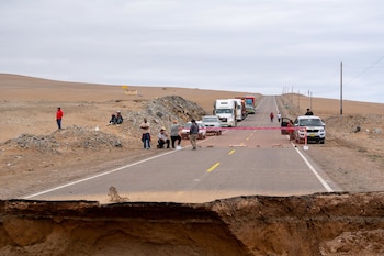 Carretera con tramo destruido por erosión de la calzada, vehículos detenidos en fila, barrera de seguridad, personas a los costados y paisaje desértico