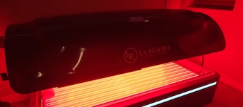 Una máquina de terapia de luz roja con una tapa oscura y tubos naranjas brillantes en una habitación iluminada de rojo. Se ve el logo 'LA RÉSERVE'