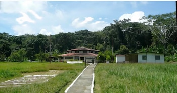 Vista panorámica de una instalación militar con un edificio principal de techo marrón, flanqueado por dos edificios pequeños, un camino de concreto y vegetación frondosa
