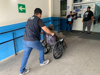 Un hombre con chaleco de PGN empuja a una persona en una silla de ruedas por una rampa de concreto en un pasillo. Una flecha verde a la izquierda