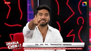 Hombre con barba y camisa blanca apunta con el dedo. Fondo oscuro con luces de neón rojas y textos de programa de televisión