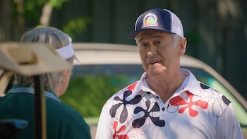 Hombre de edad media con gorra y polo estampado hablando con una persona de espaldas que viste visera y polo verde. Un carro de golf está en primer plano