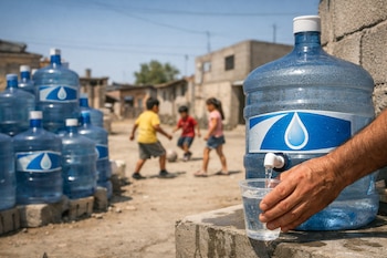 Primer plano de mano de adulto llenando vaso con agua de garrafón; en el fondo, niños juegan en una calle de tierra con casas humildes bajo un cielo azul despejado.