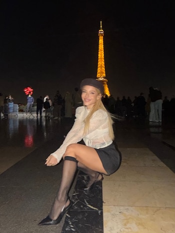 Una mujer con boina oscura, blusa blanca y falda posa agachada en un piso de baldosas de noche, con la Torre Eiffel iluminada al fondo