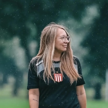 Una mujer de cabello rubio, con gafas, viste una camiseta deportiva negra con un escudo rojo y blanco mientras gotas de lluvia caen a su alrededor