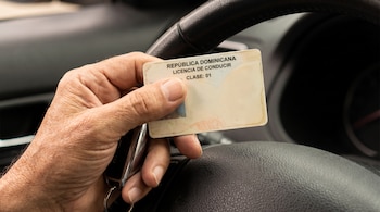 Una mano sostiene una licencia de conducir de República Dominicana y una llave metálica frente al volante de un automóvil.
