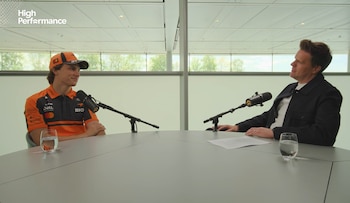 Oscar Piastri sonríe sentado a una mesa con un micrófono, vestido con gorra y polo naranja de McLaren, frente a un hombre con chaqueta oscura en un estudio