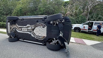 Un SUV oscuro está volcado de lado en una carretera, mostrando su parte inferior. Un vehículo de paramédicos blanco y una persona se ven en el fondo