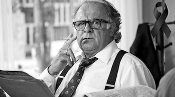 Un hombre de gafas, camisa, corbata y tirantes sostiene un cigarrillo encendido junto a un lazo negro en la esquina superior derecha.