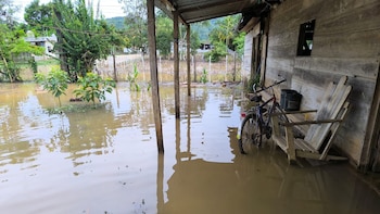 Una casa de madera con un porche inundado por agua marrón, mostrando una bicicleta y una silla de madera parcialmente sumergidas, y árboles al fondo
