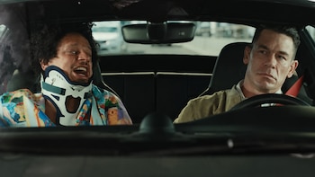 Eric André con collarín y camisa colorida en el asiento del copiloto de un coche. John Cena conduce el coche con una chaqueta beige