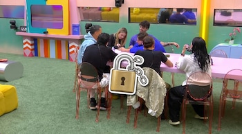 Ocho jóvenes sentados en sillas transparentes alrededor de una mesa rosa sobre alfombra verde. Hay una pantalla, reloj, cámaras y un candado dorado superpuesto.