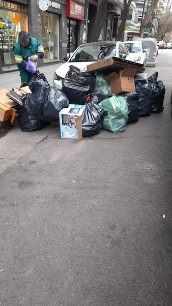 Hombre con uniforme verde y guantes morados, bolsas de basura negras y verdes, cajas de cartón, calle, acera, coche blanco, edificios, árboles, contenedores
