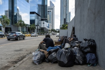 Personas sentadas en una acera junto a bolsas de basura, con rascacielos de vidrio y automóviles al fondo.