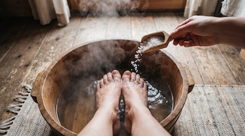 Pies sumergidos en un recipiente de madera con agua caliente y vapor, una mano vierte sal desde una cuchara, en un suelo de madera con una alfombra.