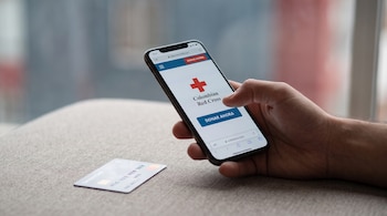 Una mano sostiene un teléfono móvil que exhibe la página web de la Cruz Roja Colombiana, mostrando su logo y el botón "Donar Ahora", junto a una tarjeta bancaria.