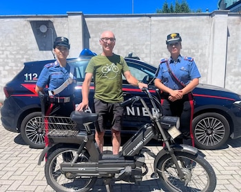 Hombre calvo en camiseta verde entre dos agentes Carabinieri con uniforme, junto a un ciclomotor GARELLI negro y un coche patrulla de la policía