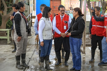 Grupo de personas con botas, algunos con chalecos rojos, de pie sobre suelo mojado y con barro. Una mujer con chaqueta negra estrecha la mano de un hombre con chaleco rojo