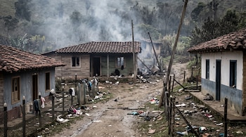 Casas de madera con ventanas rotas, un camino de tierra lleno de escombros, postes inclinados y humo en una zona rural.