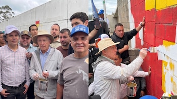 Un grupo de hombres, liderados por Álvaro Uribe, pinta un muro de bloques con secciones de pintura roja, amarilla y blanca. Uribe lleva sombrero y pincel.