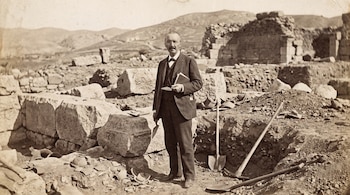 Un hombre con traje sostiene un libro y una paleta de mano en un sitio arqueológico con ruinas, bloques de piedra, fragmentos de cerámica y herramientas de excavación.