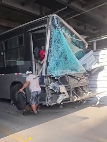Vista frontal y lateral de un autobús Metropolitano gris con el parabrisas y la parte delantera gravemente dañados. Un hombre manipula un neumático, otro está dentro