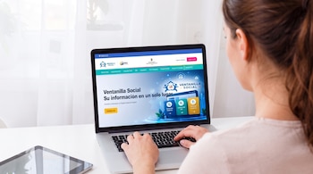Una mujer vista de espaldas escribe en una computadora portátil que muestra un sitio web gubernamental sobre una mesa blanca.