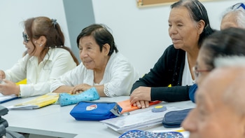 Un grupo de mujeres adultas mayores sentadas en un aula, con libretas y cartucheras sobre una mesa blanca. Una usa lentes de sol