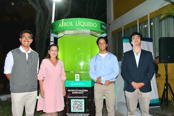 Cuatro personas, una mujer en vestido rosa y tres hombres, posan frente a un gran tanque cilíndrico transparente con líquido verde, identificado como "ÁRBOL LÍQUIDO"