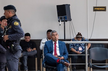 Dos agentes de policía uniformados de pie a la izquierda, un hombre en traje azul y corbata roja sentado al centro, y un letrero de prensa visible