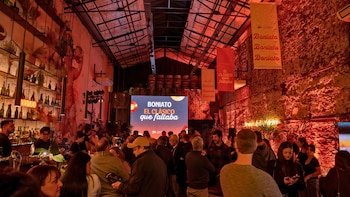 Vista interior de un evento concurrido bajo intensa luz roja; una pantalla gigante muestra 'BONIATO EL CLÁSICO que faltaba'; hay un bar y varias pancartas