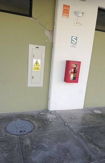 Una pared verde claro con grietas y yeso desprendido sobre un panel eléctrico, junto a una columna blanca con extintor rojo y escombros en el suelo