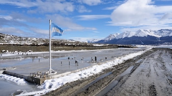 Una bandera argentina en un mástil junto a una plataforma de hormigón sobre un terreno con nieve y barro, con montañas nevadas al fondo