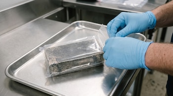 Manos enguantadas de azul sellan bolsa transparente con placa metálica gris en bandeja de acero. La bolsa tiene texto "RADIOACTIVE MATERIAL" y "DEPLETED URANIUM".