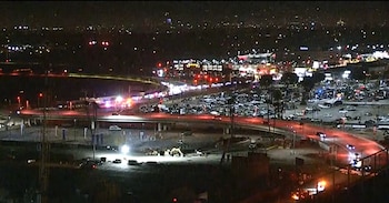 Imagen nocturna aérea de una zona urbana con carreteras, un paso elevado, luces de tráfico, vehículos de emergencia y una excavadora bajo focos intensos