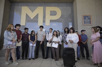 Grupo de personas, incluyendo una mujer hablando en un micrófono, está de pie frente a un edificio con letras 'MP' y una placa. Un parlante está en el suelo
