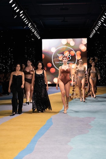 Varias modelos caminan por una pasarela con alfombra colorida, presentando lencería y atuendos. Al fondo, una pantalla muestra un logo y luces desenfocadas