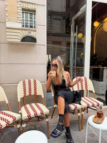 Mujer rubia con gafas de sol sentada en una silla de café, bebe de un vaso con pajita, frente a una cafetería con pasteles