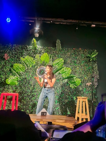 Mujer en el centro de un escenario con micrófono, pared verde con hojas, luces azules, dos sillas y público