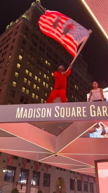 Dos hombres en lo alto de un letrero de Madison Square Garden por la noche; uno ondea una bandera de Estados Unidos