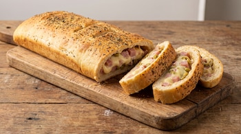 Pan relleno largo con orégano espolvoreado, cortado en rodajas sobre una tabla de madera, que expone su relleno de jamón, queso y aceitunas.