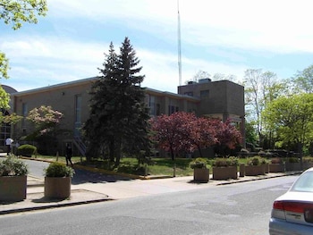 Edificio de color beige con ventanas, rodeado de árboles verdes y uno rojizo. Una calle, macetas con plantas y la parte trasera de un coche plateado en primer plano