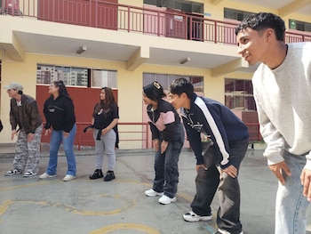 Seis jóvenes adultos, cinco mujeres y un hombre, se agachan al aire libre, mirando hacia la derecha, con un edificio amarillo y barandales rojos al fondo