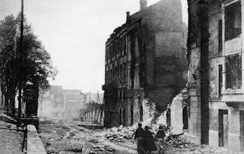 Una calle de una ciudad en blanco y negro, con edificios parcialmente derrumbados y escombros. Tres figuras pequeñas caminan entre los restos